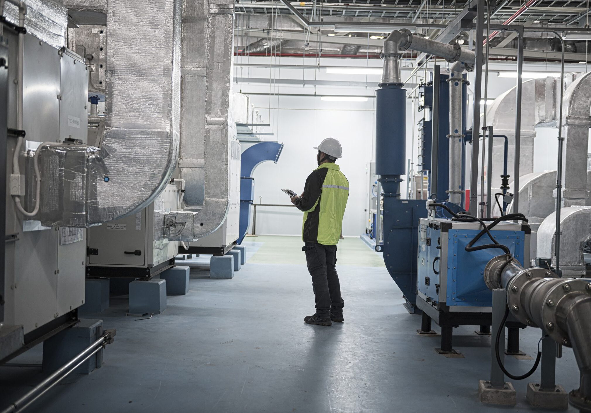 professional Asian engineer or technician in safety gear performs a routine inspection using a digital tablet inside a modern industrial facility. Surrounded by high-tech piping and equipment, he checks operational systems in a controlled, clean, and safe work environment.